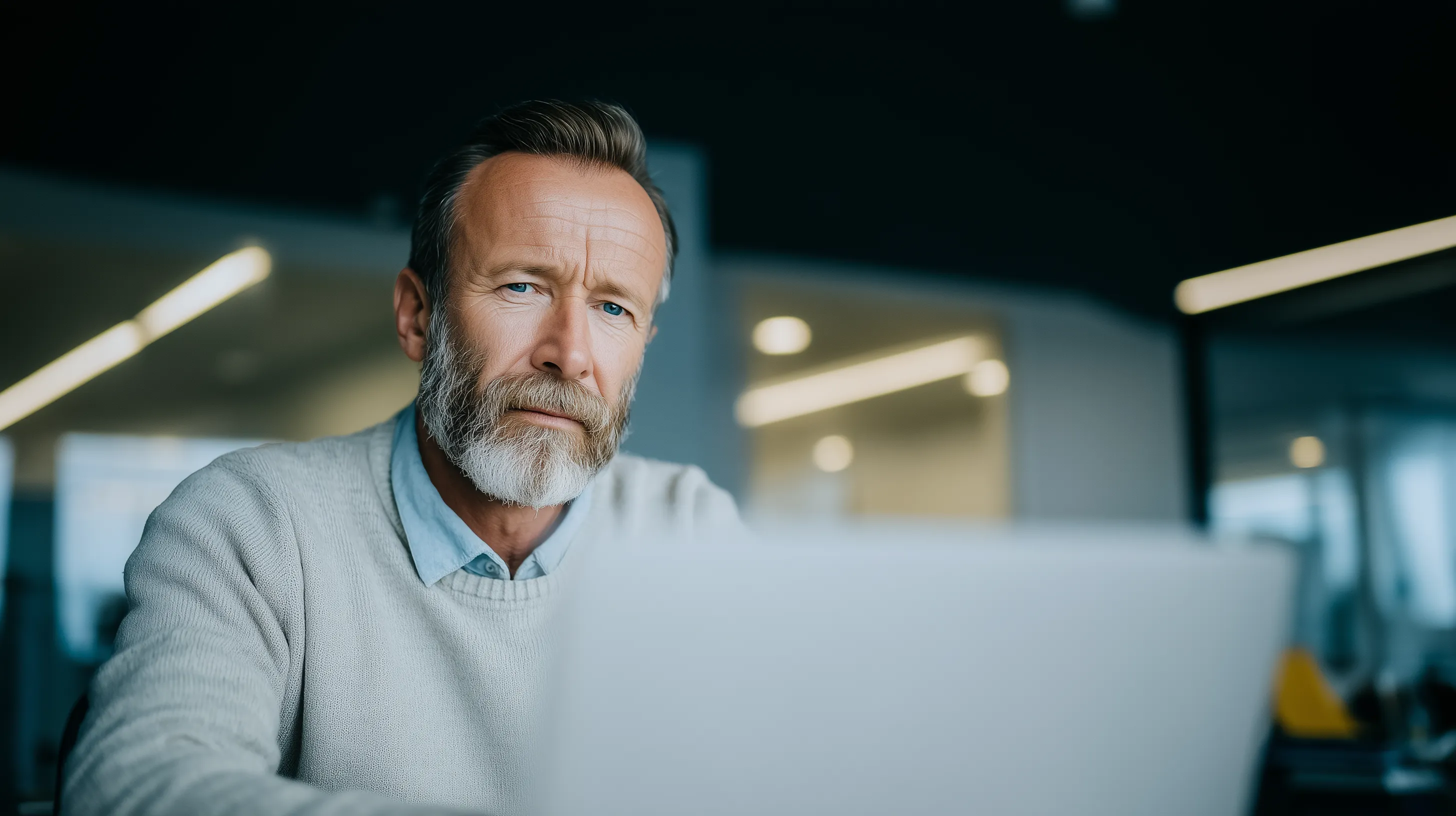 a middle-aged man with a beard in business casual clothing sits in front of a computer. His expression is thoughtful and contemplative.